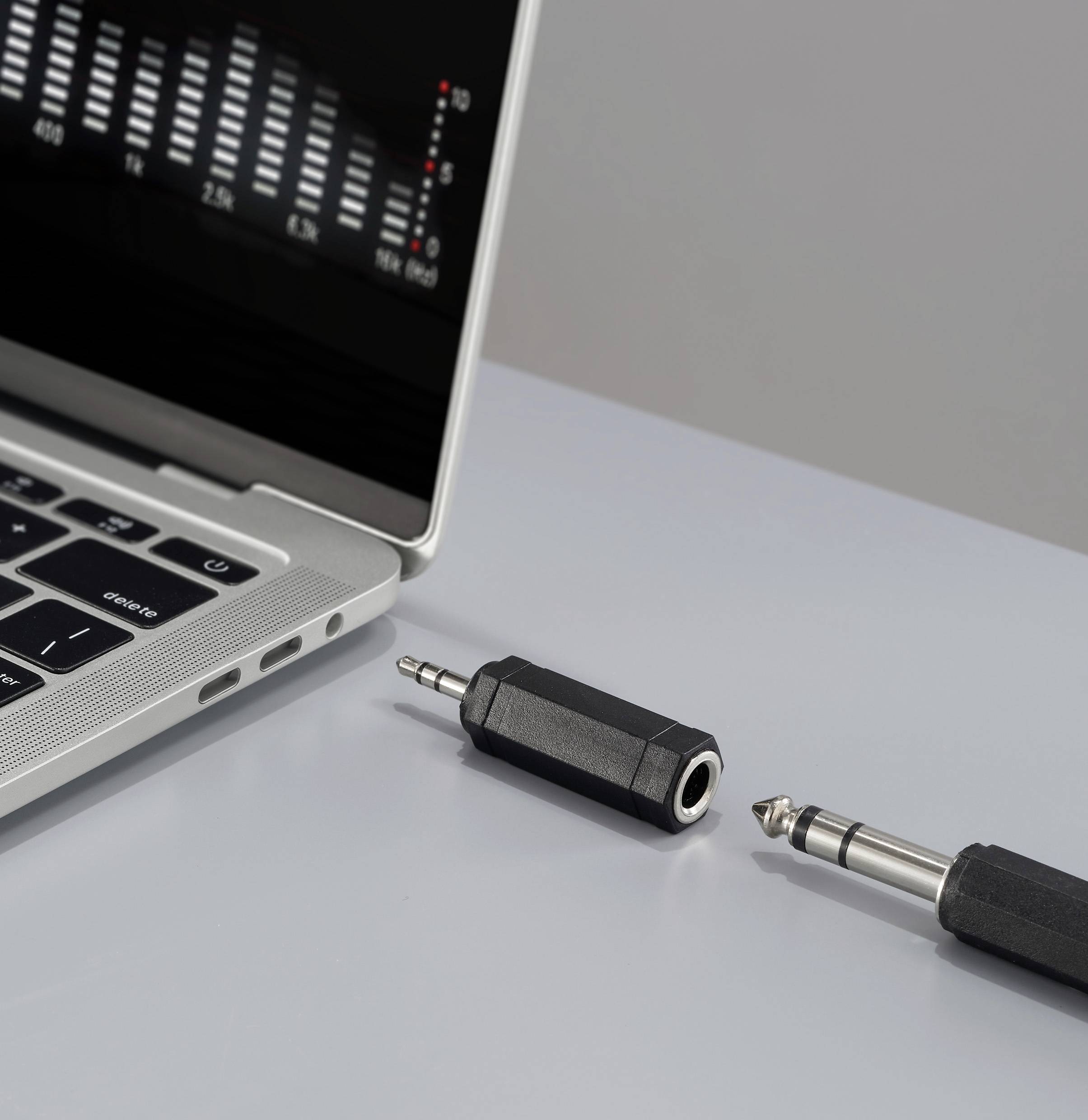 Laptop s otevřeným programem zvukového mixéru na obrazovce leží vedle 1/4 palcového audio jacku a adaptéru na šedém povrchu.
