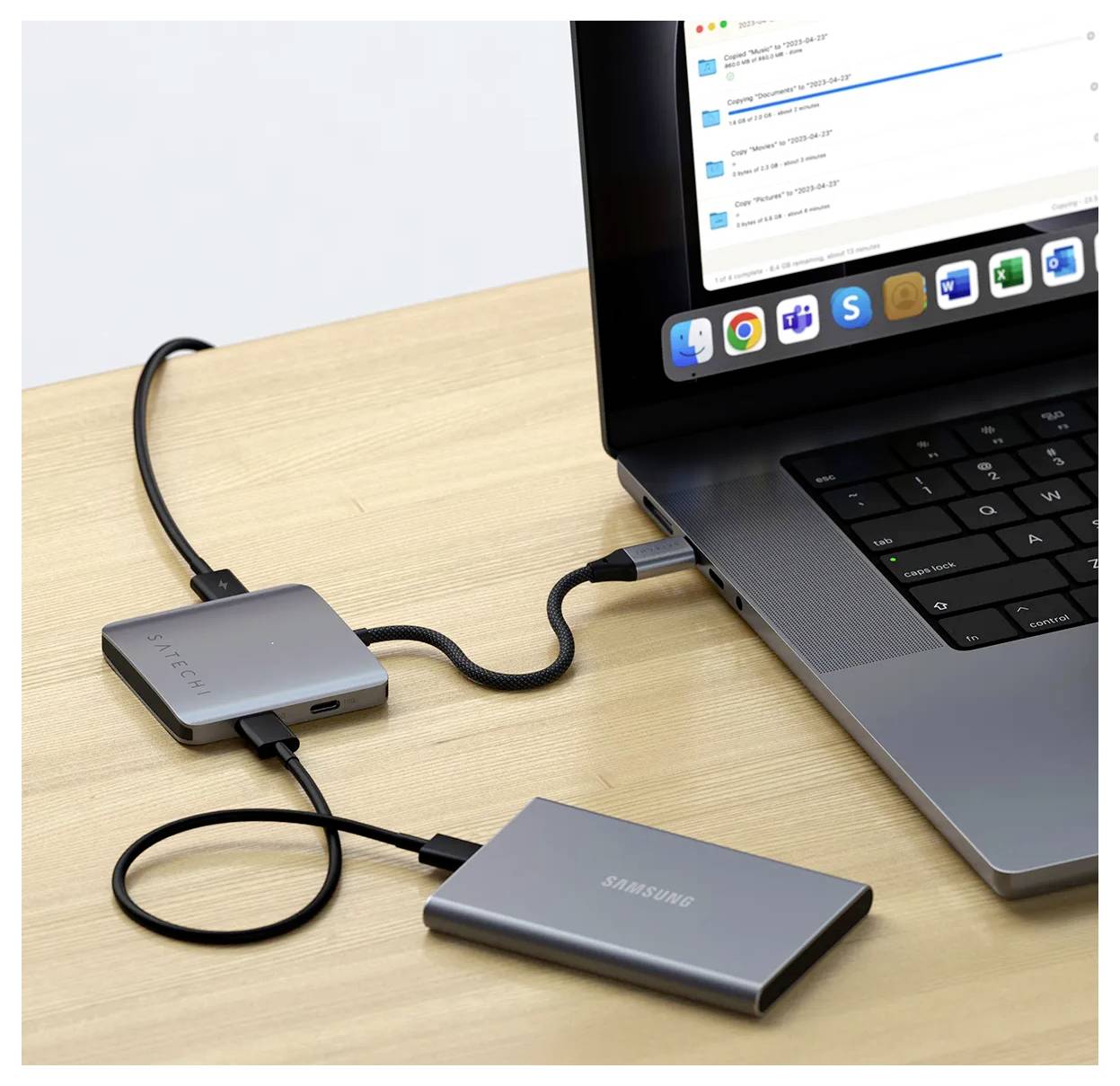 Laptop připojený k externímu pevnému disku a rozbočovači s více porty na dřevěném stole, zobrazující přenos dat nebo nastavení připojení zařízení.
