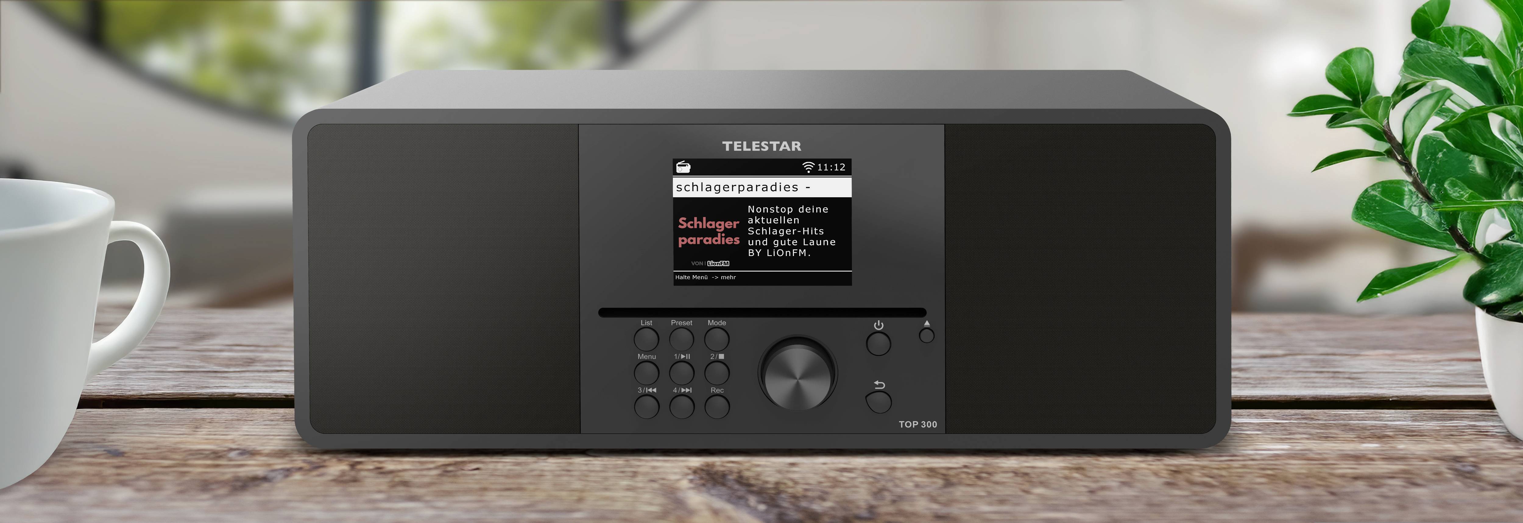 Černé digitální rádio TELESAR stojí na dřevěném stole s displejem zobrazujícím nápis 'Schlagerparadies', obklopené šálkem a výhledem z okna.