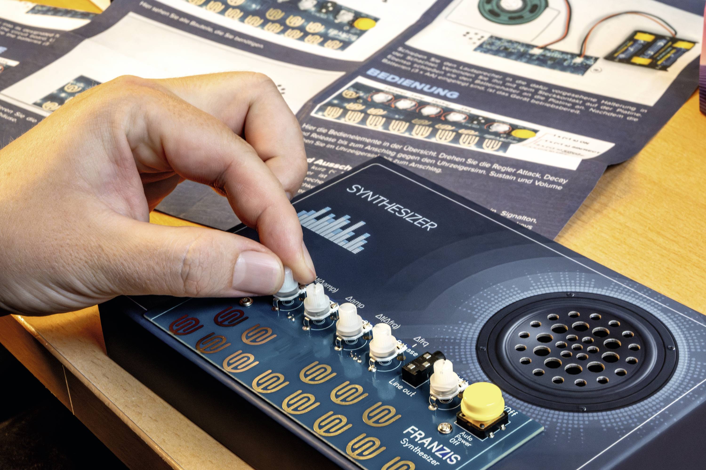 Ruka nastavuje knoflík na malé syntezátorové desce označené nápisem "SYNTHESIZER". Na stole je viditelný manuál s montážními pokyny.
