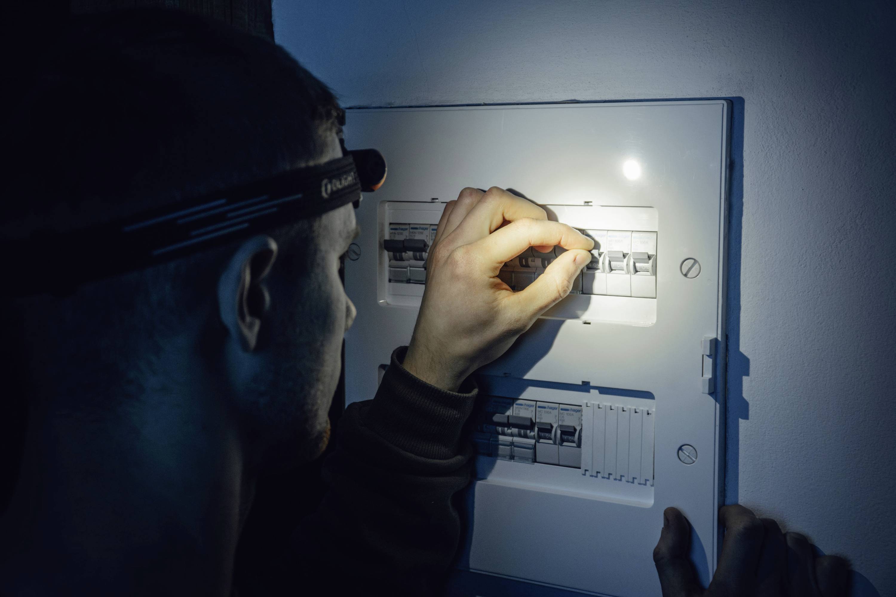 Osoba s čelovkou kontroluje elektrický rozvaděč a soustředí se na nastavení jednoho z vypínačů v nevýrazně osvětleném prostoru.