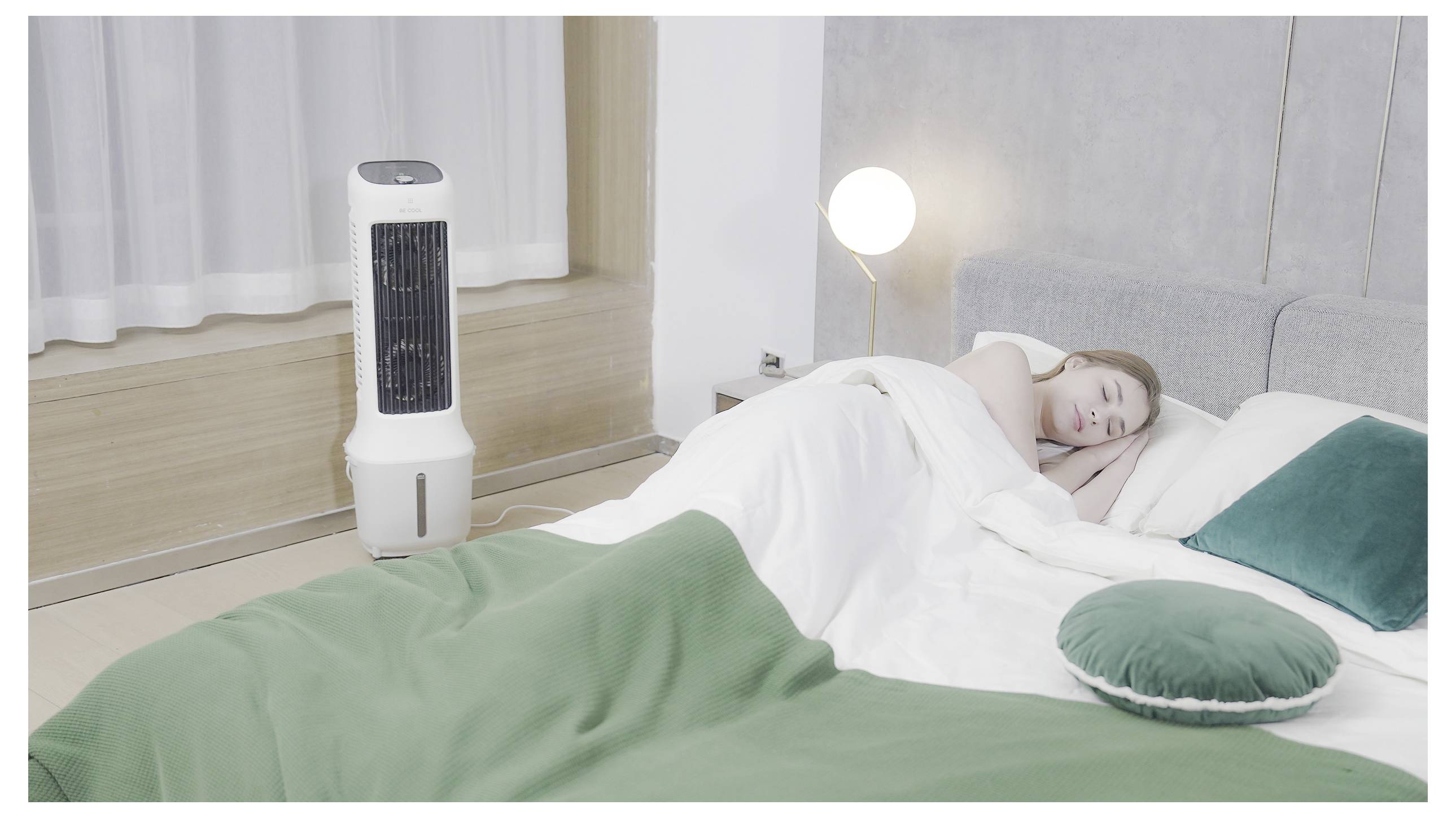 Osoba spící v moderní ložnici s bílo-zeleným motivem, vedle věžového ventilátoru a zapnuté lampy na nočním stolku.