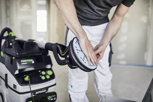Osoba obsluhuje ruční elektrickou brusku připojenou k jednotce pro odsávání prachu v místnosti, která je právě rekonstruována.