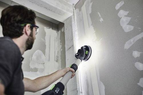 Osoba používá elektrický nástroj k broušení sádrokartonu v místnosti ve výstavbě, přičemž se zaměřuje na spoj mezi dvěma deskami.