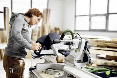 Osoba obsluhující kotoučovou pilu na dřevo ve světlé dílně, soustředěná na přesné řezání. Jsou viditelné kusy dřeva a velká okna.