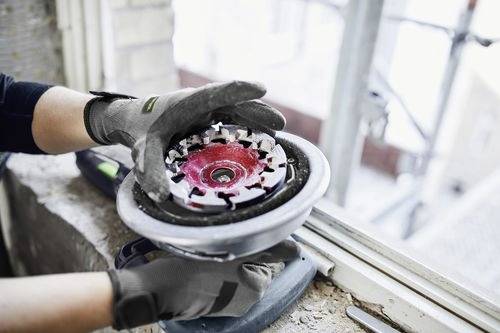 Osoba v ochranných rukavicích drží a kontroluje kruhový brusný nástroj poblíž okna, což naznačuje prostředí stavby nebo renovace.