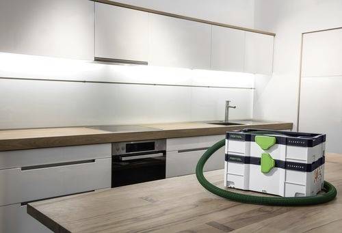 Moderní kuchyně s elegantními bílými skříňkami a dřevěnými pracovními deskami. Na ostrůvku stojí kompaktní vysavač s viditelnými stočenými hadicemi.