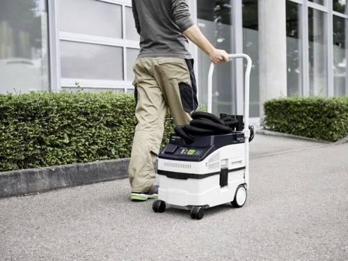 Osoba tlačí mobilní vysavač po chodníku vedle budovy s velkými okny a zastřiženými keři.