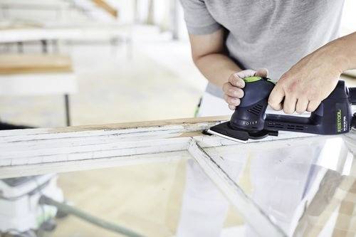 Osoba používá ruční elektrickou brusku k vyhlazení hrany dřevěného rámu, pravděpodobně v přípravě na malování nebo renovaci.