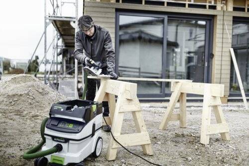 Osoba v čepici a rukavicích používá elektrický nástroj na kus dřeva podepřený koz(l)íky na staveništi.