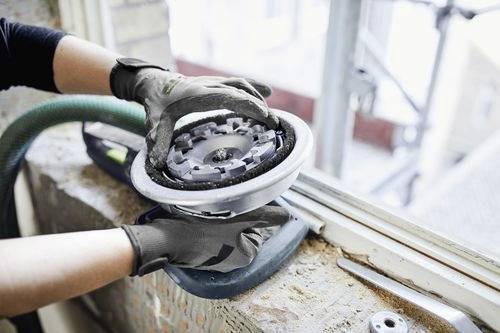 Dělník v rukavicích používá elektrickou brusku na okenním rámu, vyhlazuje povrch v blízkosti staveniště.