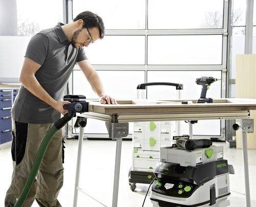Osoba v ochranných brýlích brousí desku pomocí elektrického nástroje v dílně. Odsavač prachu je připojen, což zajišťuje čisté pracovní prostředí.