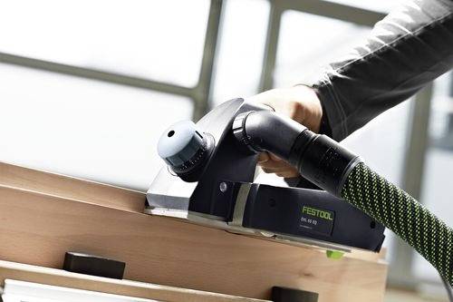 Osoba používá ruční elektrický hoblík Festool k vyhlazení hrany dřevěné desky uvnitř místnosti.