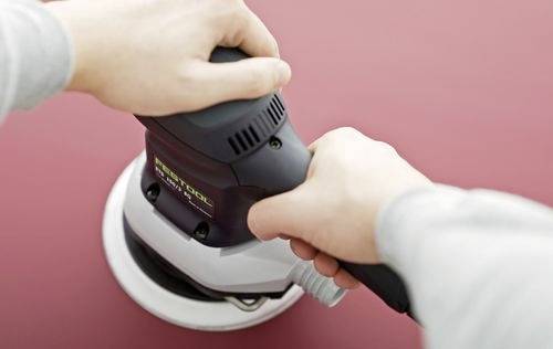 Ruce drží ruční elektrickou brusku, která vyhazuje rovný červený povrch. Zaměření je na techniku aplikace broušení.