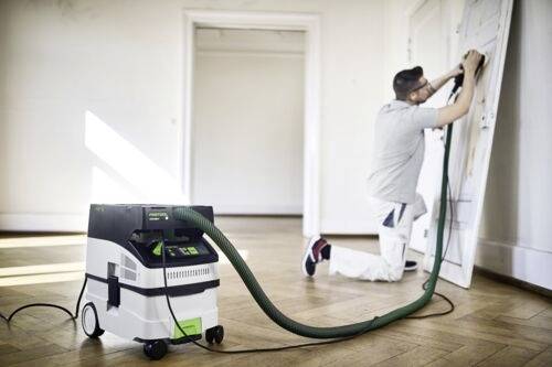 Osoba v pracovním oblečení brousí dveře elektrickým bruska připojenou k vysavači prachu na dřevěné podlaze v jasně osvětlené místnosti.
