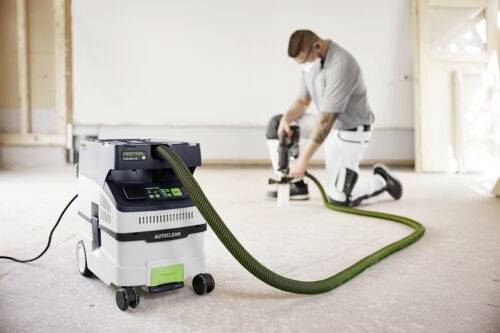 Dělník na stavbě klečí na podlaze, má nasazenou ochrannou roušku a používá elektrický nástroj připojený k vysavači pro odsávání prachu v místnosti.