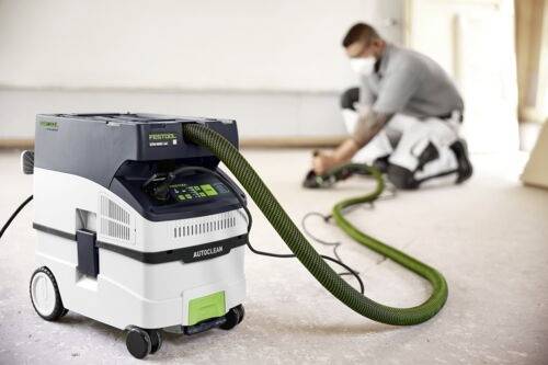 Osoba v masce klečí na podlaze a přebrušuje dřevo, připojená k vysavači Festool v popředí pro čistější práci.