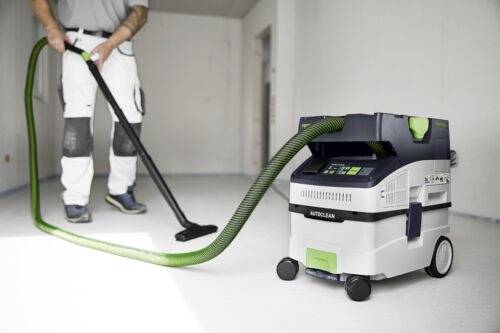 Osoba oblečená v pracovních kalhotách ovládá kompaktní, kolečkový vysavač označený 'Autoclean' pomocí zelené hadice v bílé místnosti.