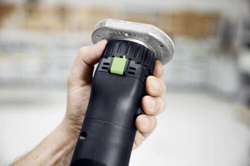 Ruka osoby držící černou a zelenou elektrickou ruční brusku s kovovým povrchem v prostředí dílny.
