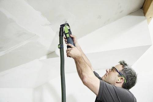 Osoba v ochranných brýlích používá ruční elektrickou brusku na stropě, vyhlazuje povrch, přičemž je k nástroji připojena hadice.