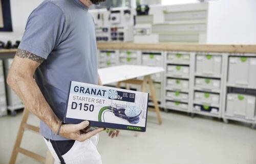 Osoba v dílně držící krabici od sady 'Granat Starter Set D150', v pozadí s úložnými regály a pracovním stolem.