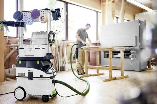 Dílna s osobou, která brousí dřevo u stolu. V popředí je mobilní odsavač prachu a stanice pro broušení, která ukazuje nástroje a vybavení.