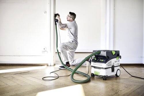 Osoba klečící při používání elektrického nářadí připojeného k přenosnému vysavači v jasně osvětlené místnosti s parketovou podlahou.