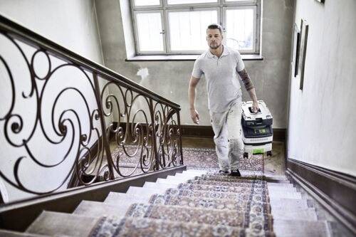 Osoba v pracovním oblečení nese kufřík s elektrickým nářadím, zatímco stoupá po kobercem potažených schodech v budově s ozdobným zábradlím a velkým oknem.