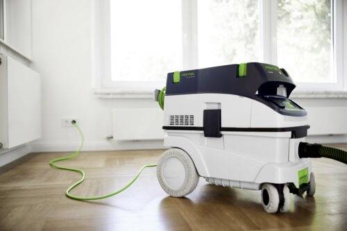 Bílý a zelený vysavač stojí na dřevěné podlaze, zapojený do elektrické zásuvky. Slunečním světlem prosvítají velká okna v pozadí.