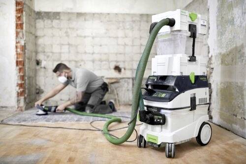 'Dělník používá elektrický nástroj na podlaze, zatímco v popředí stojí odsavač prachu připojený hadicí v částečně zrekonstruované místnosti.'