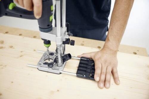 Osoba používá příklepovou vrtačku s nastavitelným vodítkem na dřevěné desce a předvádí přesnou vrtací techniku.