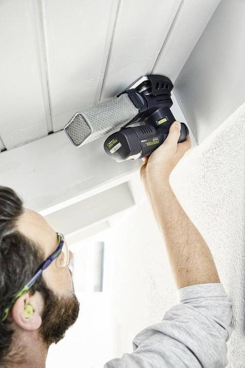 Osoba používá elektrickou brusku na stropním trámu a pro bezpečnost má nasazené ochranné brýle a špunty do uší.