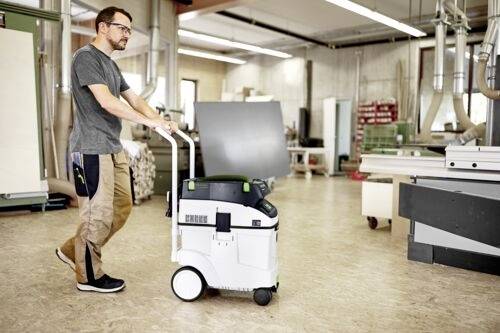 Osoba tlačí mobilní vysavač v dílně s různými nástroji a materiály kolem, což naznačuje probíhající úklid.