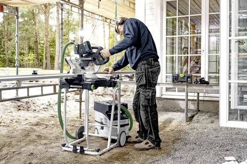 Osoba používající motorovou pilu na stavebním místě, oblečená v ochranných pomůckách. Pracuje na dřevěných trámech venku u budovy.