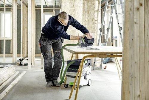 Osoba v pracovním oblečení obsluhuje elektrický nástroj na dřevěném stole v nedokončené místnosti, což naznačuje probíhající stavební projekt.