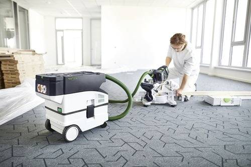 Osoba používající elektrický nástroj k řezání podlahového materiálu v jasné, nezařízené místnosti, s odsávačem prachu poblíž.
