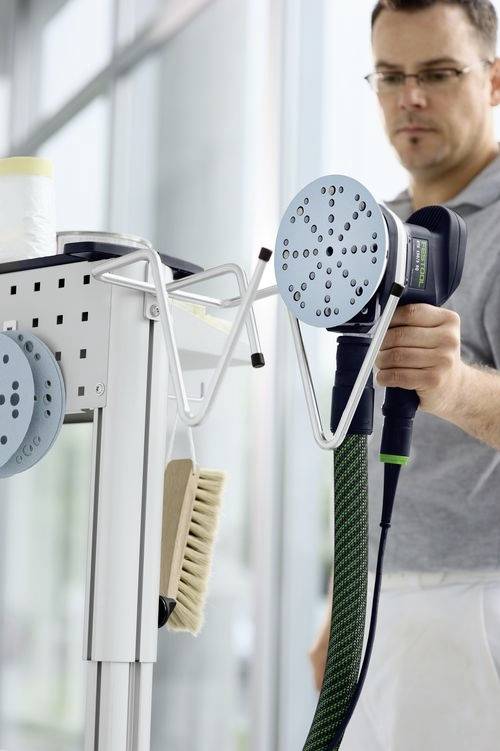 Muž používající velký elektrický nástroj s kulatou hlavou, připevněný k vozíku s různými čisticími prostředky, v jasném, moderním prostoru.