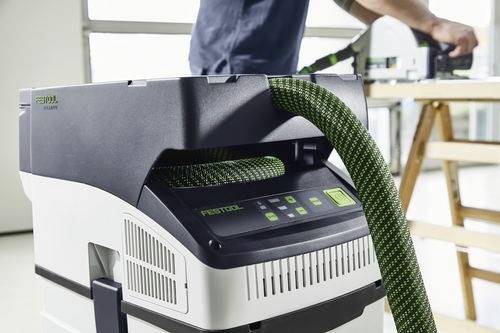 Osoba obsluhuje odsavač prachu Festool v dílně, přičemž zvýrazňuje ovládací panel a zelenou hadici na přístroji.