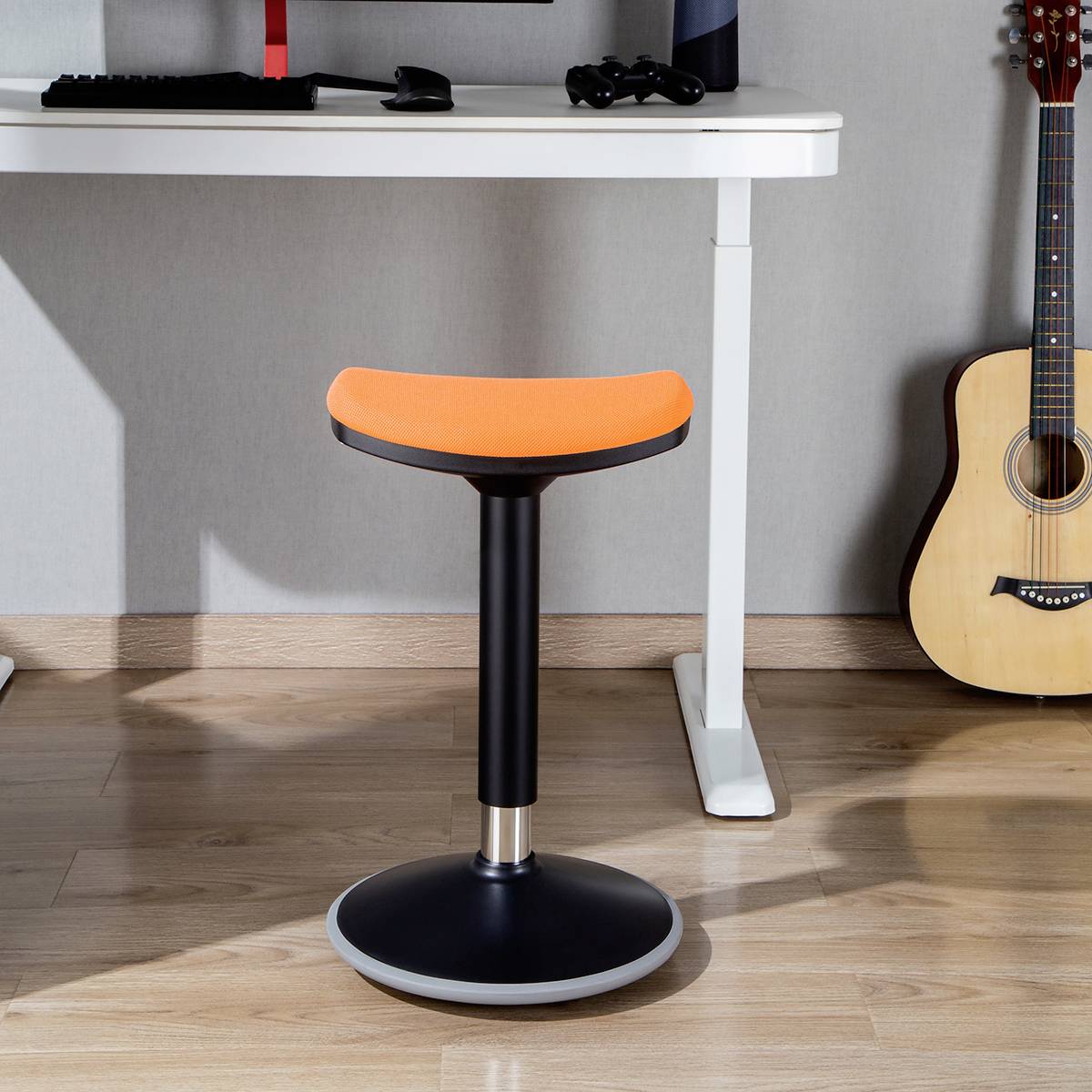 Oranžová stolička stojí na dřevěné podlaze před bílým stolem s počítačovým monitorem. Akustická kytara se opírá o zeď.