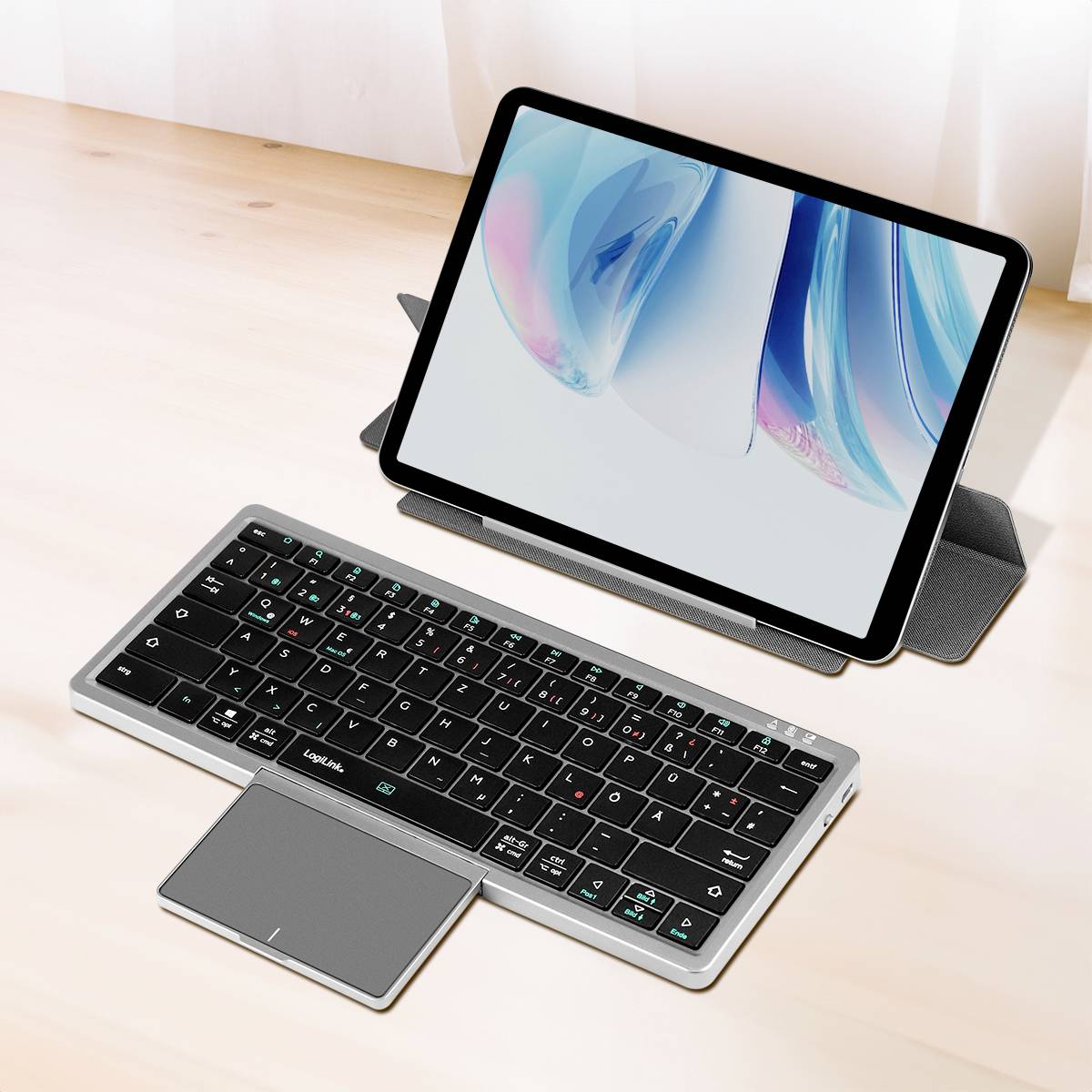 Tablet stojí opřený na pouzdře za bezdrátovou klávesnicí s touchpadem na dřevěném stole a zobrazuje lehký abstraktní design na obrazovce.