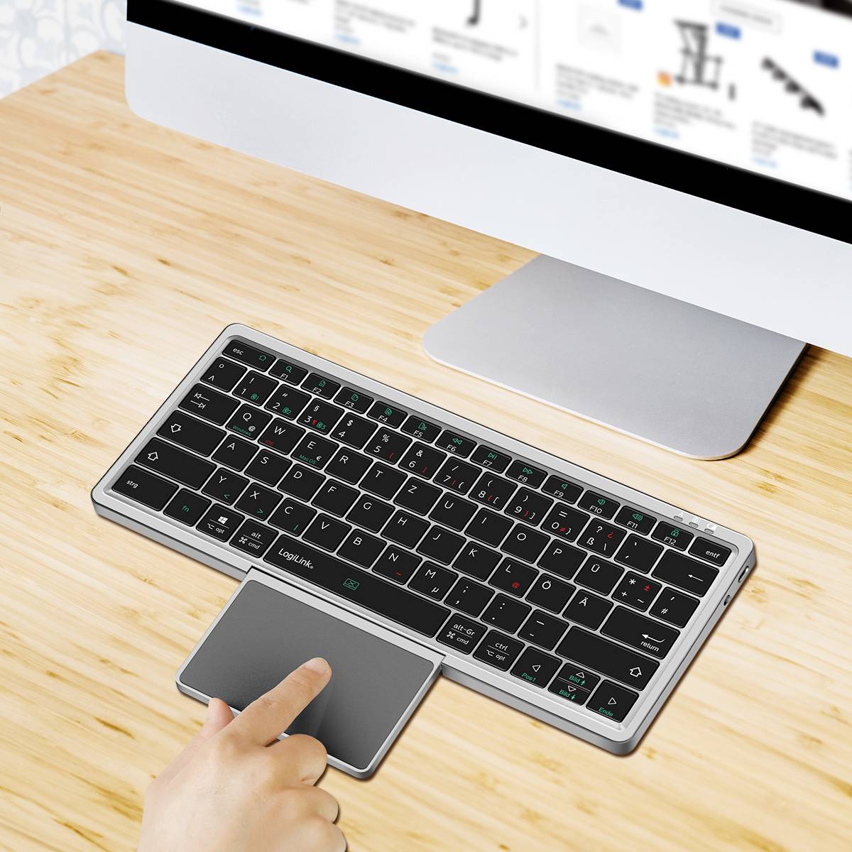Ruka interagující s digitálním touchpadem vedle bezdrátové klávesnice na dřevěném stole; v pozadí je počítačový monitor.