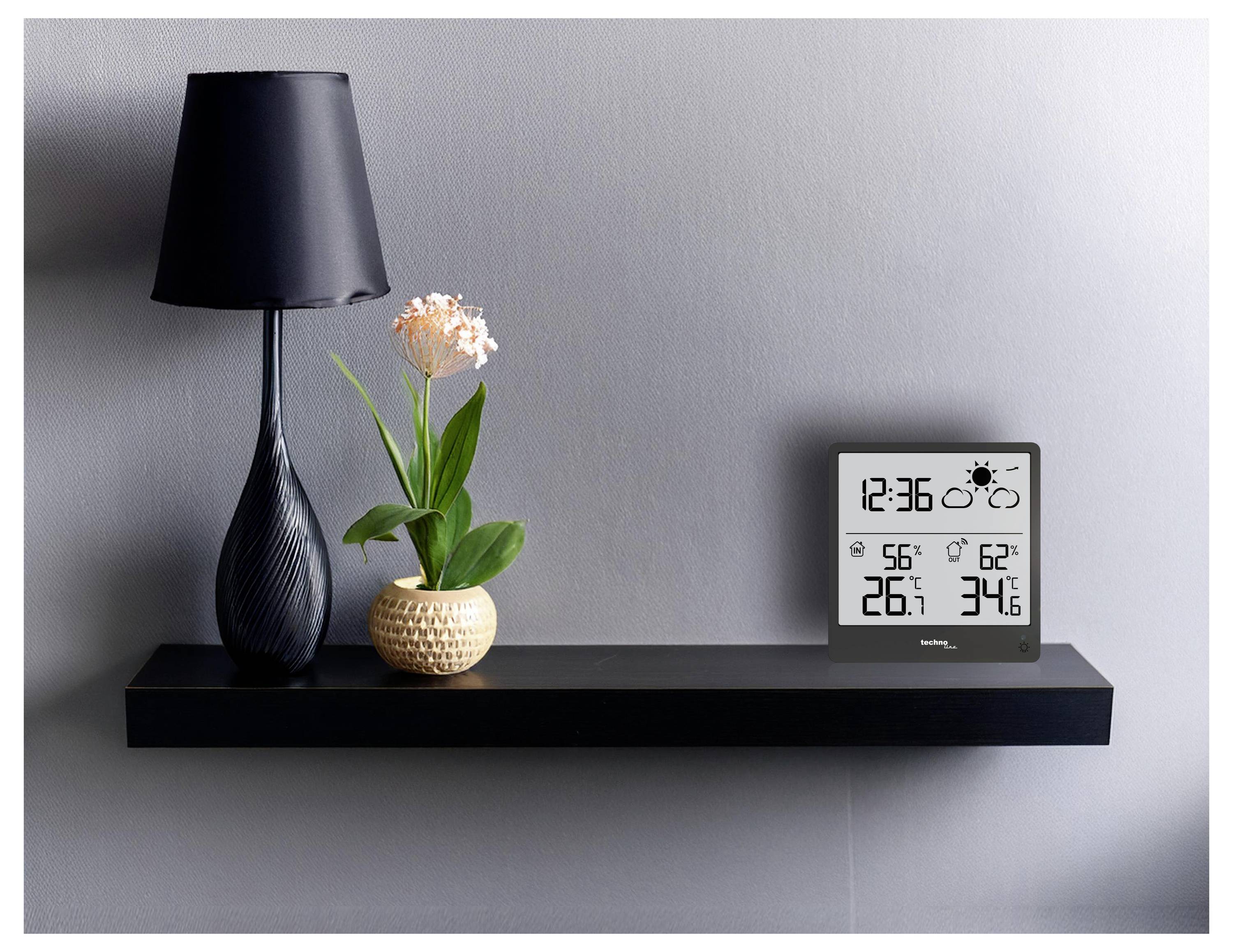 Černá police s černou lampou, malým pokojovým květináčem a digitální meteorologickou stanicí zobrazující čas, teplotu a meteorologické ikony.