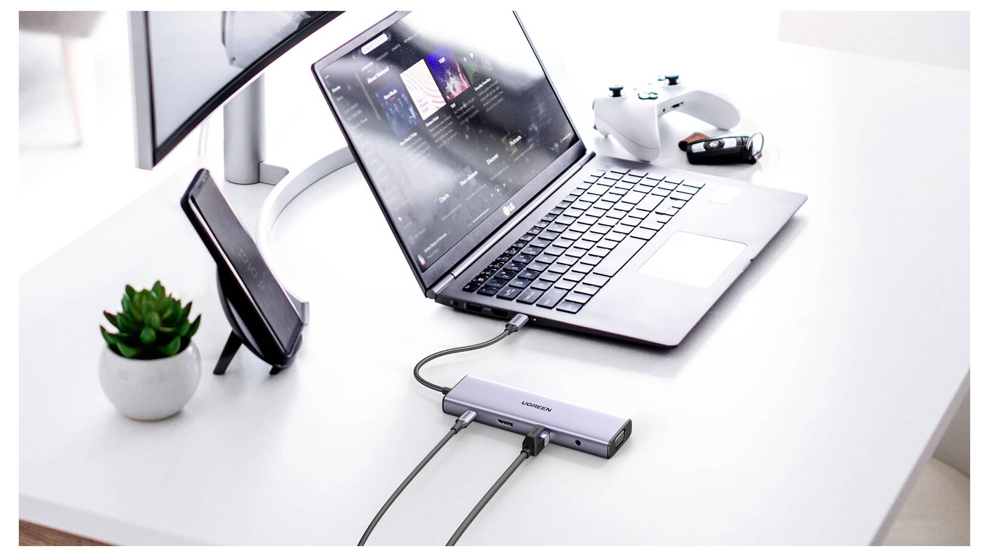 Laptop s otevřenou hudební aplikací, připojený k dokovací stanici na bílém stole s monitorem, dokovací stanicí na smartphone, ovladačem Xbox a rostlinou.