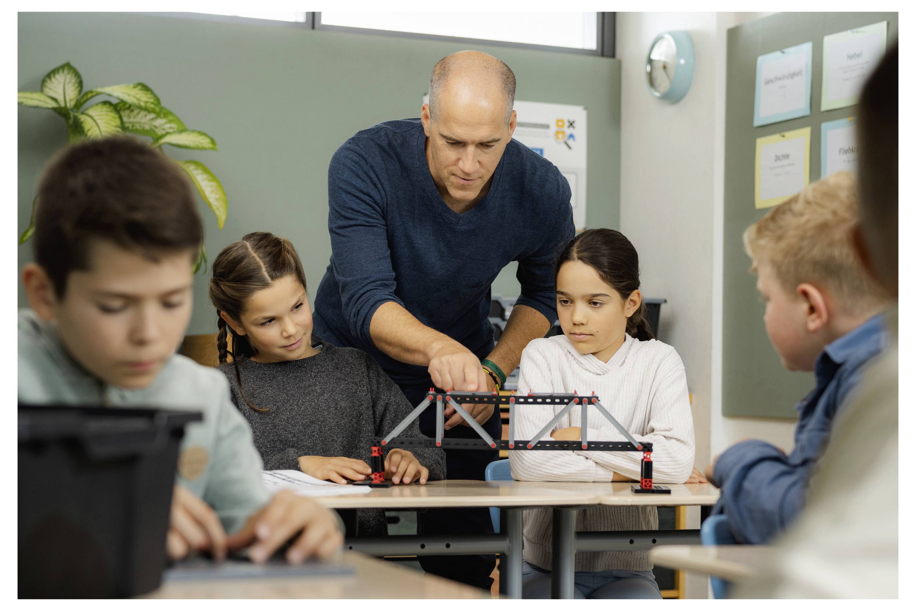 Učitelka předvádí stavbu modelu mostu čtyřem pozorným studentům ve třídě, čímž podporuje praktické učení a spolupráci.