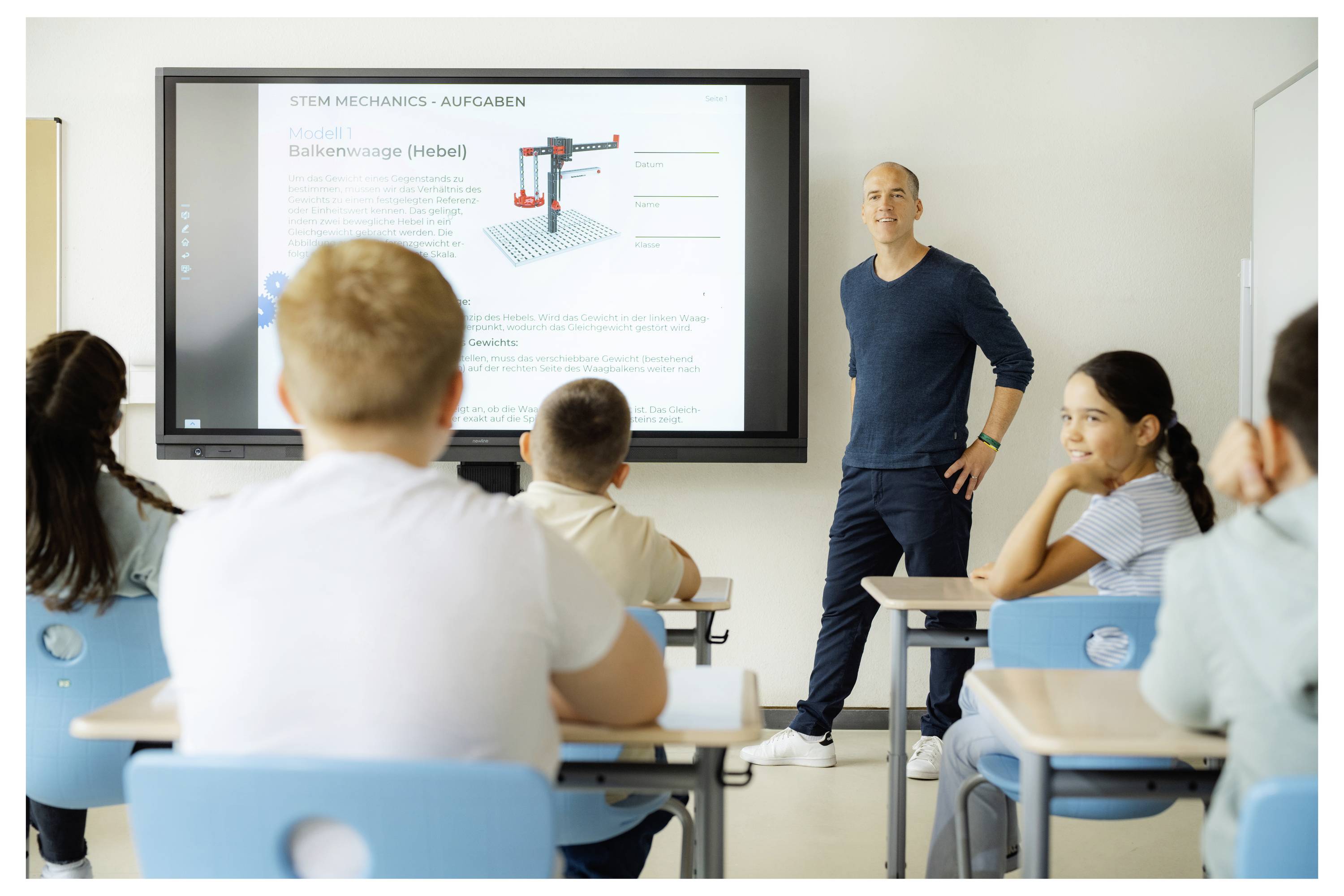 Učitel stojí vedle obrazovky zobrazující výuku 'STEM Mechaniky - Úlohy', zatímco mladí studenti sedící u lavic pozorně naslouchají.