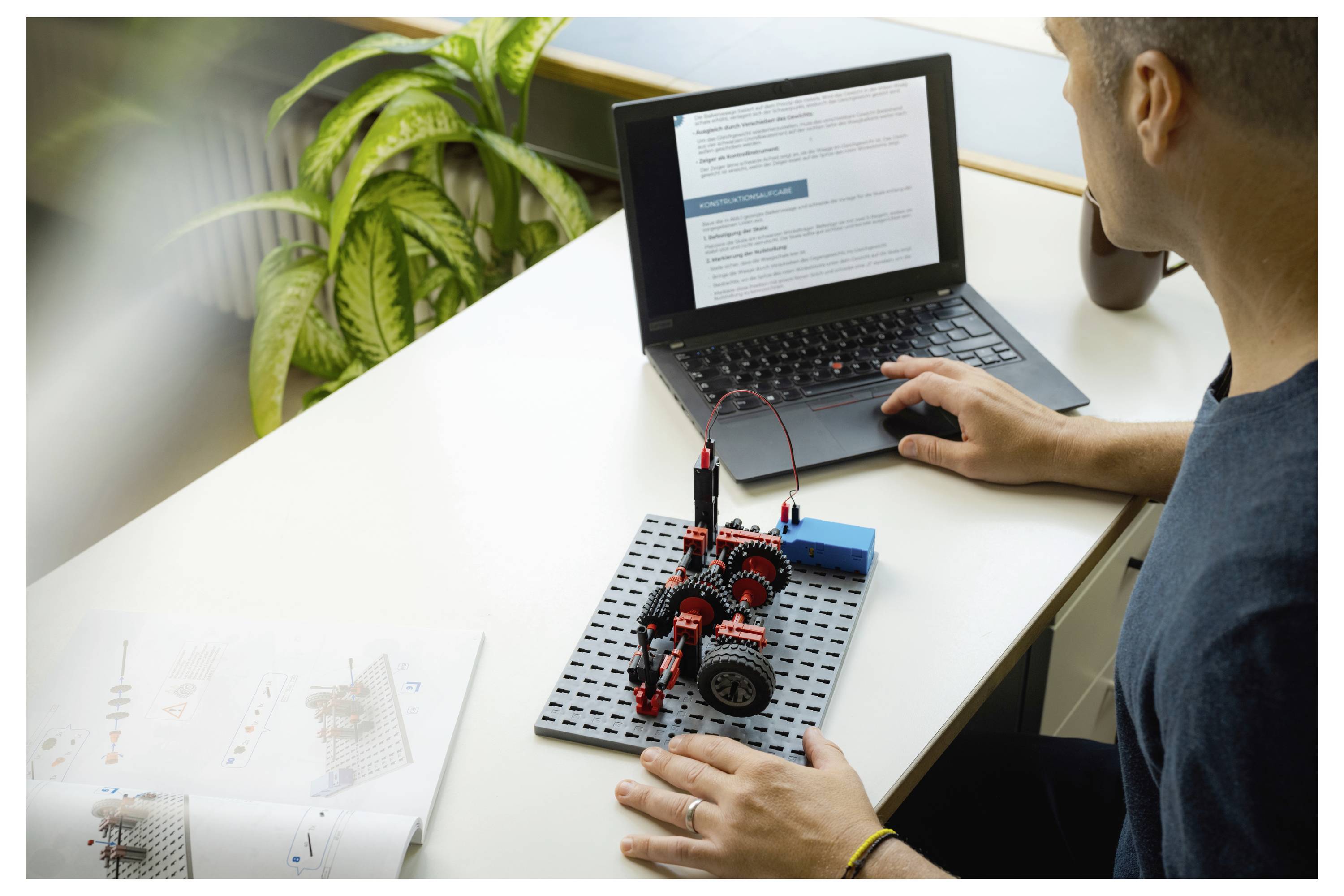 Osoba sestavuje robotickou stavebnici podle pokynů zobrazených na notebooku. Pracovní prostor zahrnuje rostlinu a manuál.
