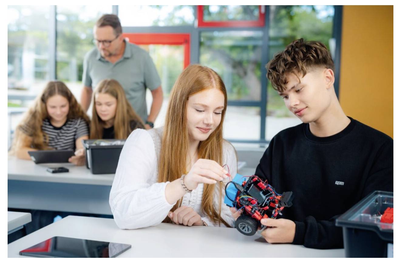 Studenti společně konstruují malého robota ve třídě, přičemž učitel a spolužáci pracují v pozadí.