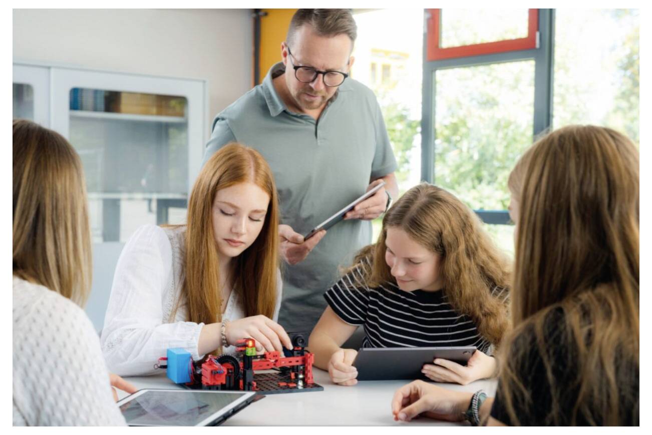 Učitel vede žáky při práci na robotickém projektu, přičemž používají tablety a model robota v světlé učebně.