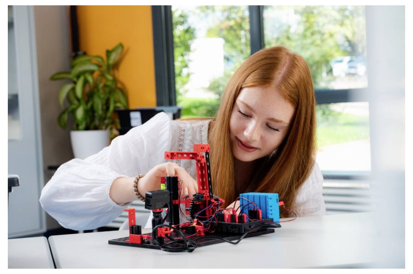 Mladá žena s dlouhými rudými vlasy pracuje na malém robotickém projektu s různými červenými a černými komponenty a vodiči na stole.