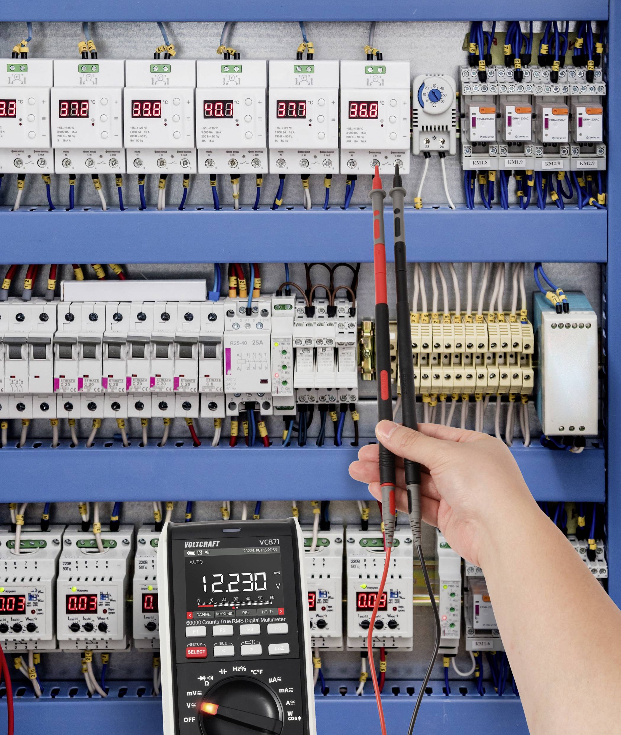 Ruka osoby drží sondy multimetru, které měří elektrický panel, s různými digitálními displeji zobrazujícími hodnoty kolem 380 voltů.